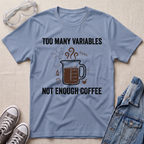 A light blue graphic t-shirt laid flat on a beige fabric background, surrounded by styling props including safety goggles, gray sneakers, and a denim jacket. The t-shirt features a dark brown coffee mug graphic with steam and the text 'TOO MANY VARIABLES NOT ENOUGH COFFEE'.