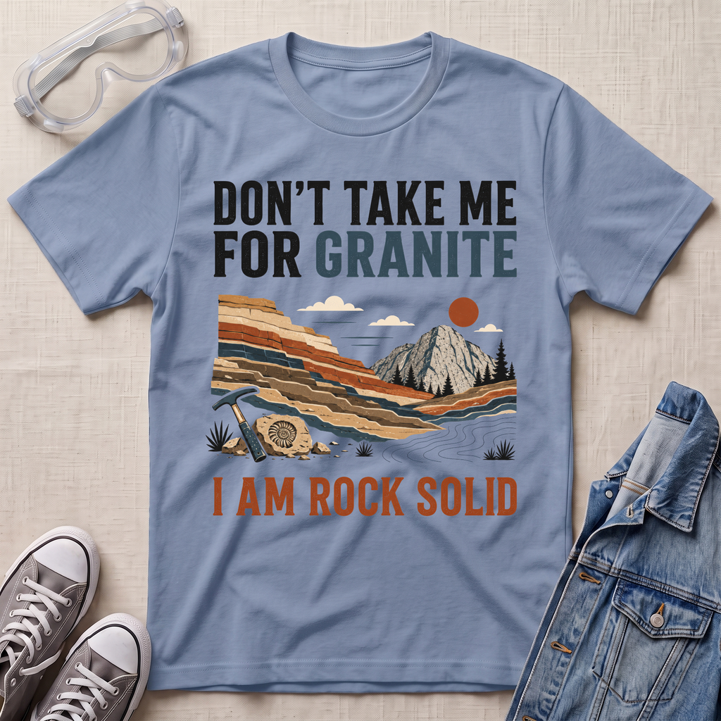 A light blue graphic t-shirt laid flat on a beige textured background, surrounded by styled props including clear safety glasses, gray sneakers, and a denim jacket. The t-shirt features a retro-style landscape graphic of a mountain and layered rock formations, with the text 'DON'T TAKE ME FOR GRANITE' above and 'I AM ROCK SOLID' below.