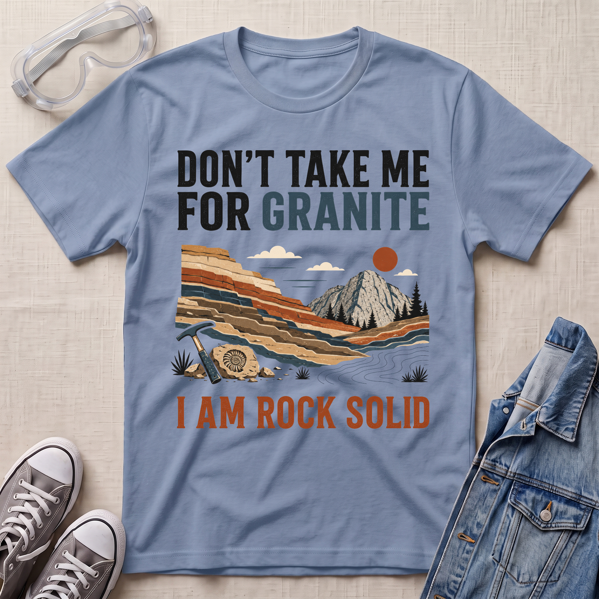A light blue graphic t-shirt laid flat on a beige textured background, surrounded by styled props including clear safety glasses, gray sneakers, and a denim jacket. The t-shirt features a retro-style landscape graphic of a mountain and layered rock formations, with the text 'DON'T TAKE ME FOR GRANITE' above and 'I AM ROCK SOLID' below.