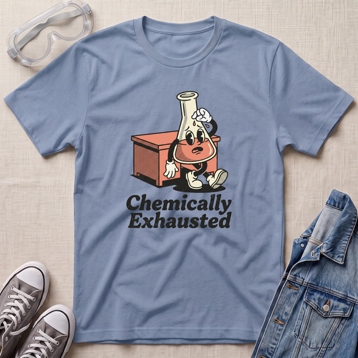 A light blue crewneck t-shirt laid flat on a beige textured background, styled with safety goggles, gray sneakers, and a denim jacket. The t-shirt features a cartoon graphic of a lab flask character with sunglasses and a sweatshirt, sitting next to a beaker, with the text 'Chemically Exhausted' printed below.