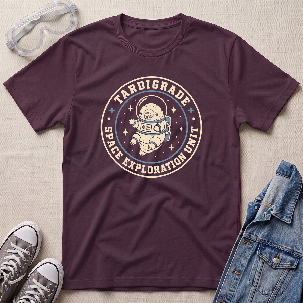 A maroon t-shirt with a retro circular graphic reading 'TARDIGRADE SPACE EXPLORATION UNIT' around an astronaut illustration, styled in a flat lay with safety goggles, gray sneakers, and a denim jacket on a beige fabric background.