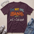 A dark plum purple graphic t-shirt laid flat on a beige fabric background, surrounded by styling props including safety glasses, gray sneakers, and a denim jacket. The t-shirt features a colorful text graphic reading 'I'M NOT ONE TO BRAGG BUT.