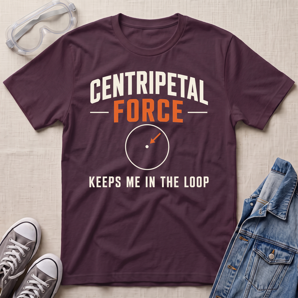 A maroon graphic t-shirt laid flat on a light beige fabric background, styled with safety goggles, gray sneakers, and a denim jacket. The t-shirt features the text 'CENTRIPETAL FORCE KEEPS ME IN THE LOOP' with a simple vector graphic in the center.