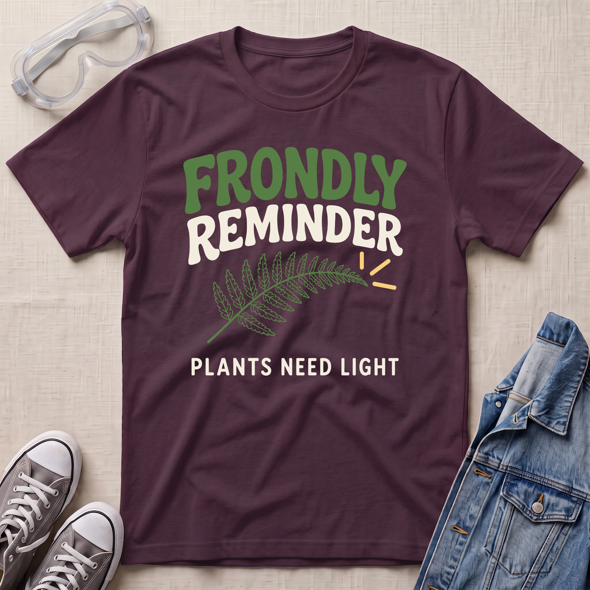 A maroon graphic t-shirt laid flat on a beige fabric background, surrounded by styled props including safety goggles, gray sneakers, and a denim jacket. The t-shirt features a green fern leaf graphic and the text 'FRONDLY REMINDER PLANTS NEED LIGHT' in green and white.