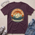 A dark plum purple t-shirt with a retro circular graphic of a smiling fish and the text 'BATHYPELAGIC ZONE' and 'Keep it Deep, Dark, and High Pressure'. The shirt is styled in a flat-lay arrangement with safety goggles, gray sneakers, and a denim jacket on a textured beige fabric background.