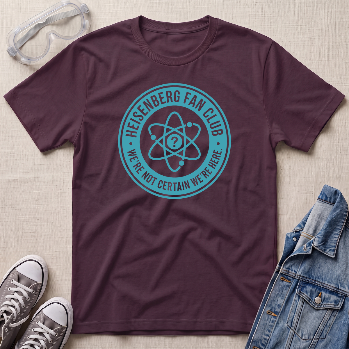 A dark plum purple t-shirt with a teal circular Heisenberg fan club graphic, styled in a flat lay with safety goggles, gray sneakers, and a denim jacket on a beige textured background.