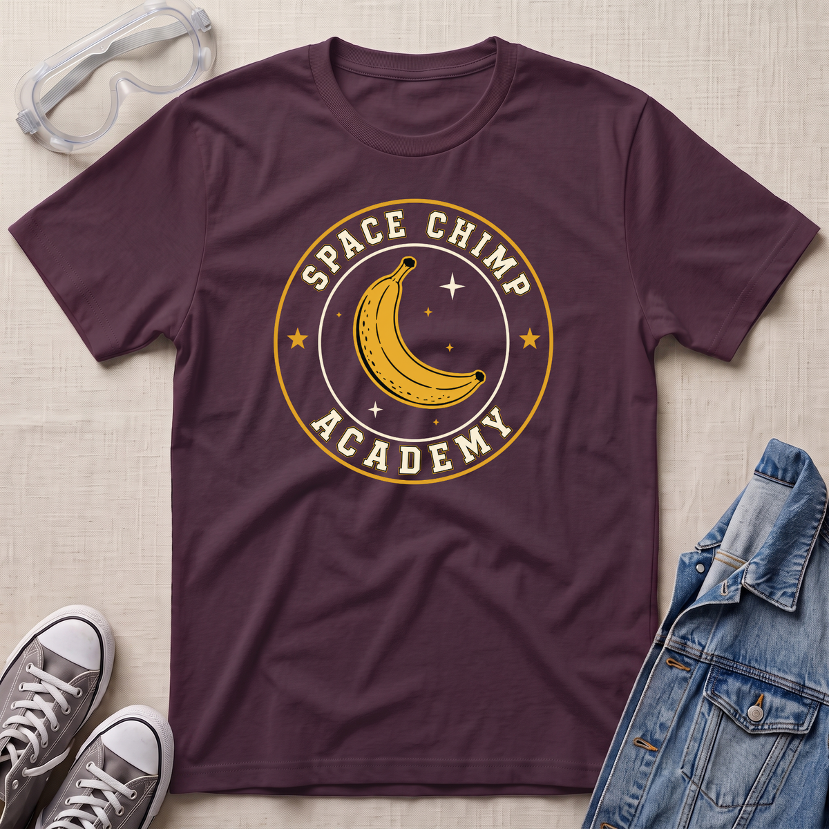 A maroon t-shirt with a circular 'Space Chimp Academy' graphic featuring a yellow banana and stars, styled in a flat lay with safety goggles, jeans, sneakers, and a denim jacket on a textured beige background.