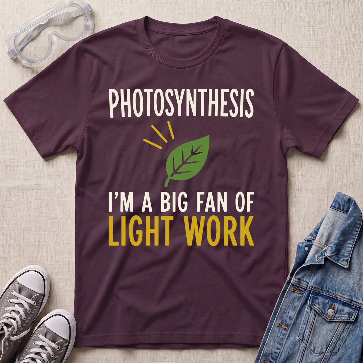 A dark plum purple graphic t-shirt laid flat on a beige fabric background, surrounded by props including safety goggles, gray sneakers, and a denim jacket. The t-shirt features a white and yellow graphic that reads 'PHOTOSYNTHESIS I'M A BIG FAN OF LIGHT WORK' with a green leaf illustration.