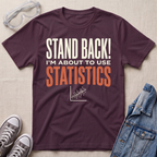 A maroon graphic t-shirt laid flat on a light beige textured background, styled with safety goggles, gray sneakers, and a denim jacket. The t-shirt features the text 'STAND BACK! I'M ABOUT TO USE STATISTICS' in cream and orange lettering, with a small white scatterplot graphic below.