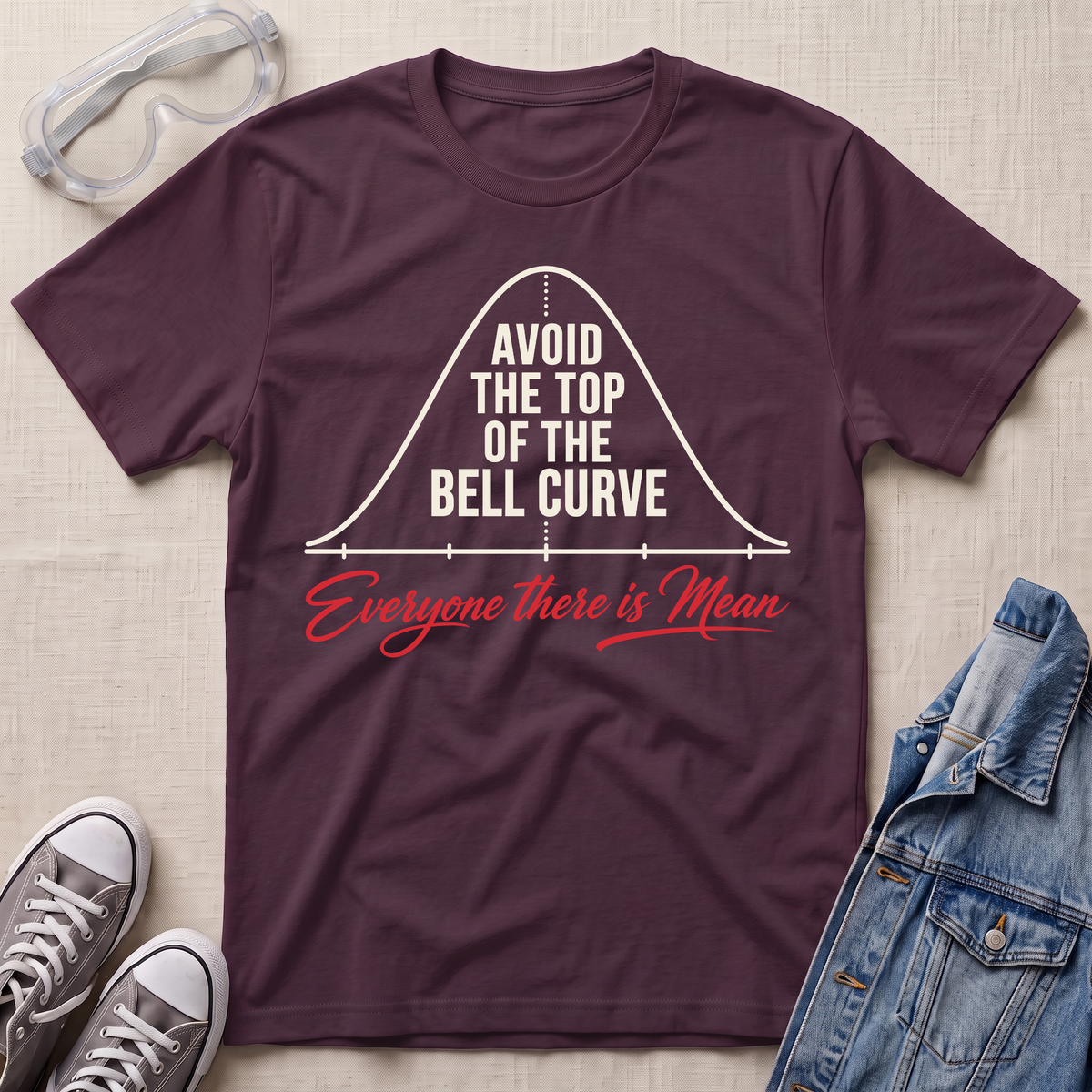 A dark plum purple graphic t-shirt laid flat on a light beige textured background, styled with safety glasses, gray sneakers, and a denim jacket. The t-shirt features a white bell curve graphic with the text 'AVOID THE TOP OF THE BELL CURVE' and 'Everyone there is Mean' in red script.