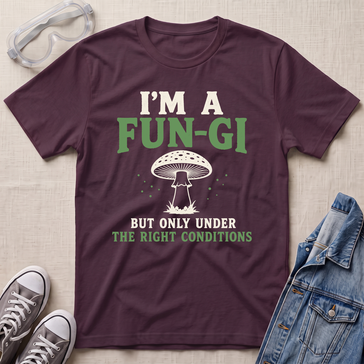 A maroon graphic t-shirt laid flat on a light beige fabric background, styled with safety goggles, gray sneakers, and a denim jacket. The t-shirt features a white and green graphic of a mushroom with the text 'I'M A FUN-GI BUT ONLY UNDER THE RIGHT CONDITIONS'.