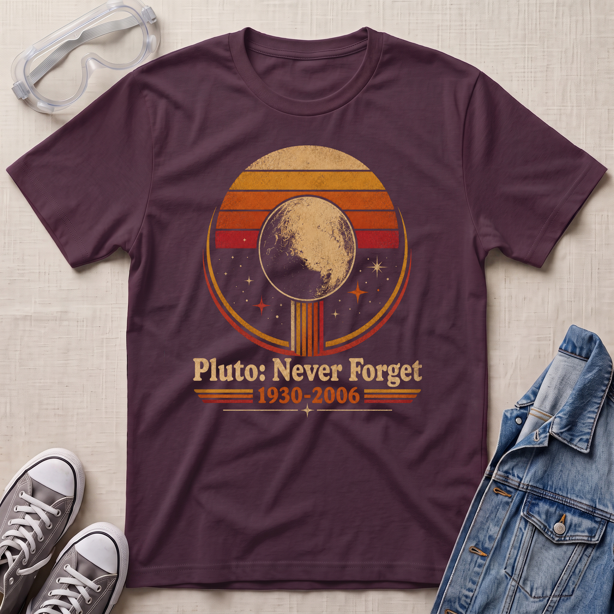 A maroon t-shirt with a retro circular graphic of Pluto, styled in a flat-lay with safety glasses, gray sneakers, and a denim jacket on a beige fabric background.