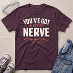 A dark plum purple graphic t-shirt laid flat on a light beige textured background, surrounded by styling props including clear safety goggles, gray sneakers, and a blue denim jacket. The t-shirt features the text 'YOU'VE GOT A LOT OF NERVE' in white and orange lettering with an orange branch graphic underneath.