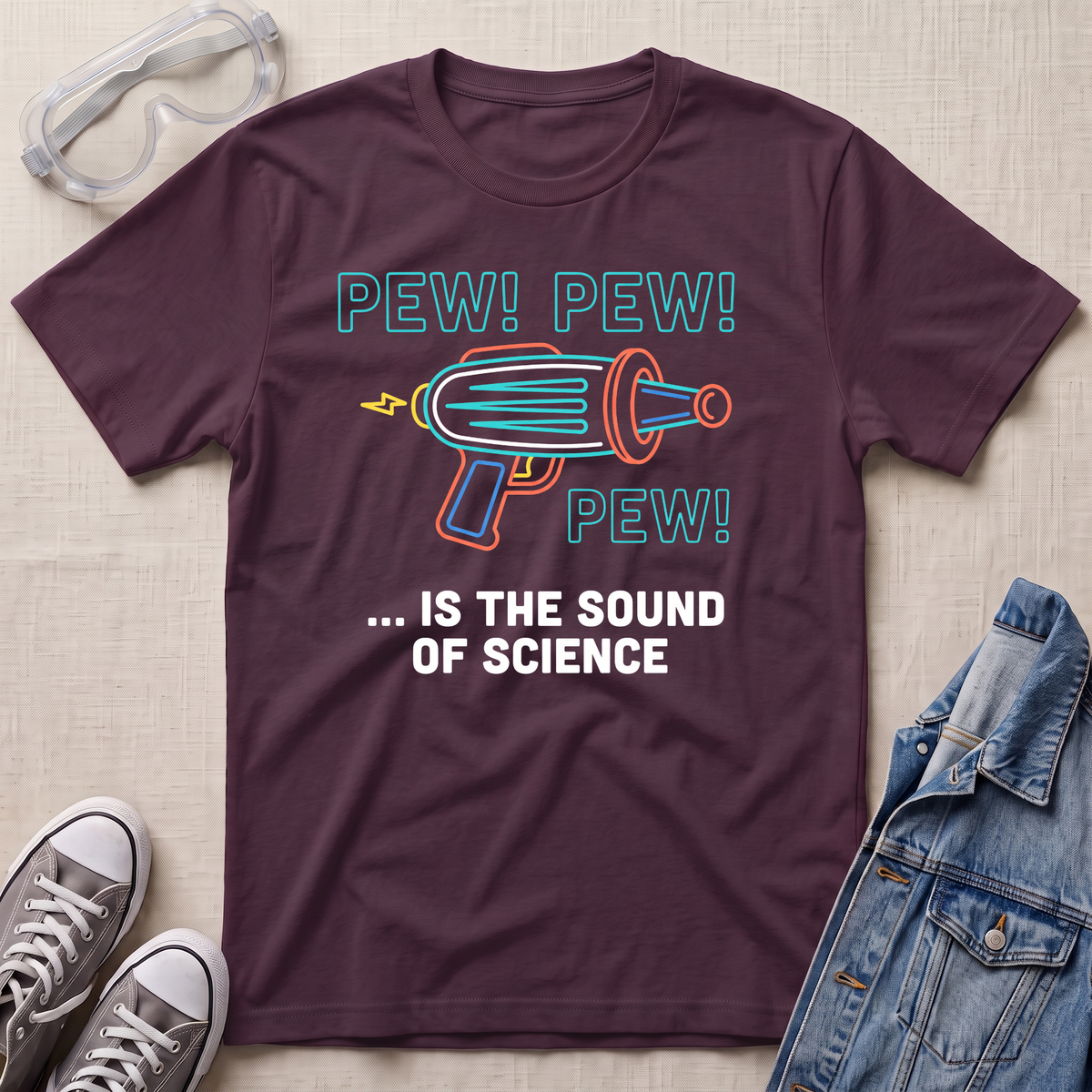 A maroon graphic t-shirt laid flat on a light beige fabric background, styled with safety goggles, gray sneakers, and a denim jacket. The t-shirt features a colorful retro toy gun illustration with the text 'PEW! PEW! PEW! ... IS THE SOUND OF SCIENCE.'