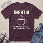 A maroon graphic t-shirt laid flat on a beige fabric background, surrounded by styling props including safety goggles, gray sneakers, and a denim jacket. The t-shirt features a white graphic of a steaming coffee mug with the text 'INERTIA REMAINING AT REST UNTIL THE CAFFEINE KICKS IN'.
