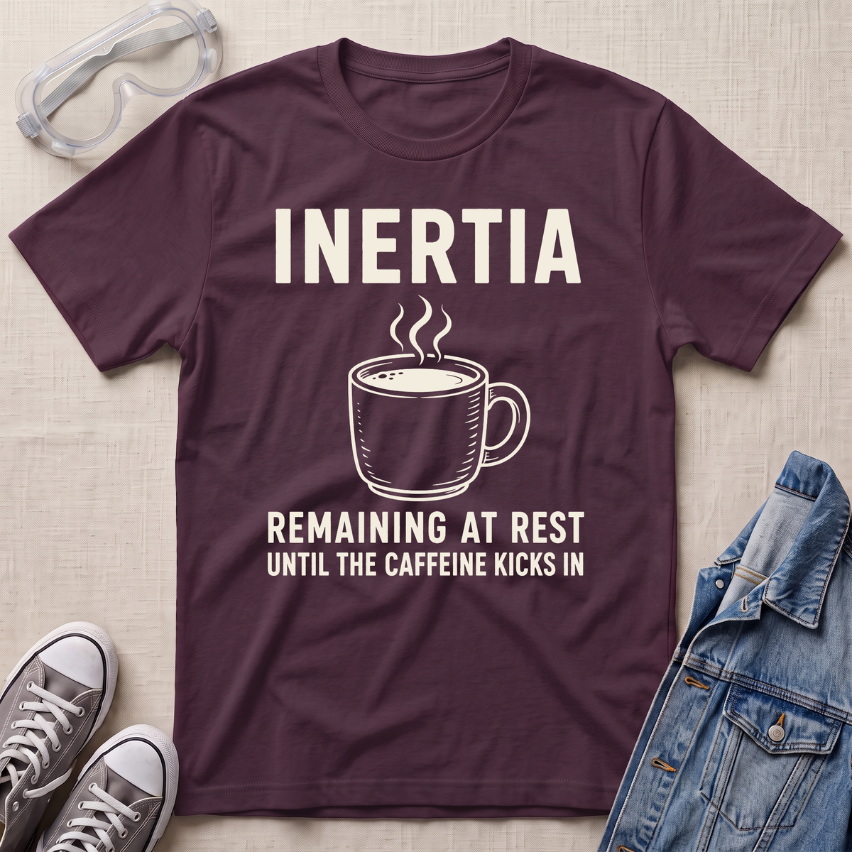 A maroon graphic t-shirt laid flat on a beige fabric background, surrounded by styling props including safety goggles, gray sneakers, and a denim jacket. The t-shirt features a white graphic of a steaming coffee mug with the text 'INERTIA REMAINING AT REST UNTIL THE CAFFEINE KICKS IN'.