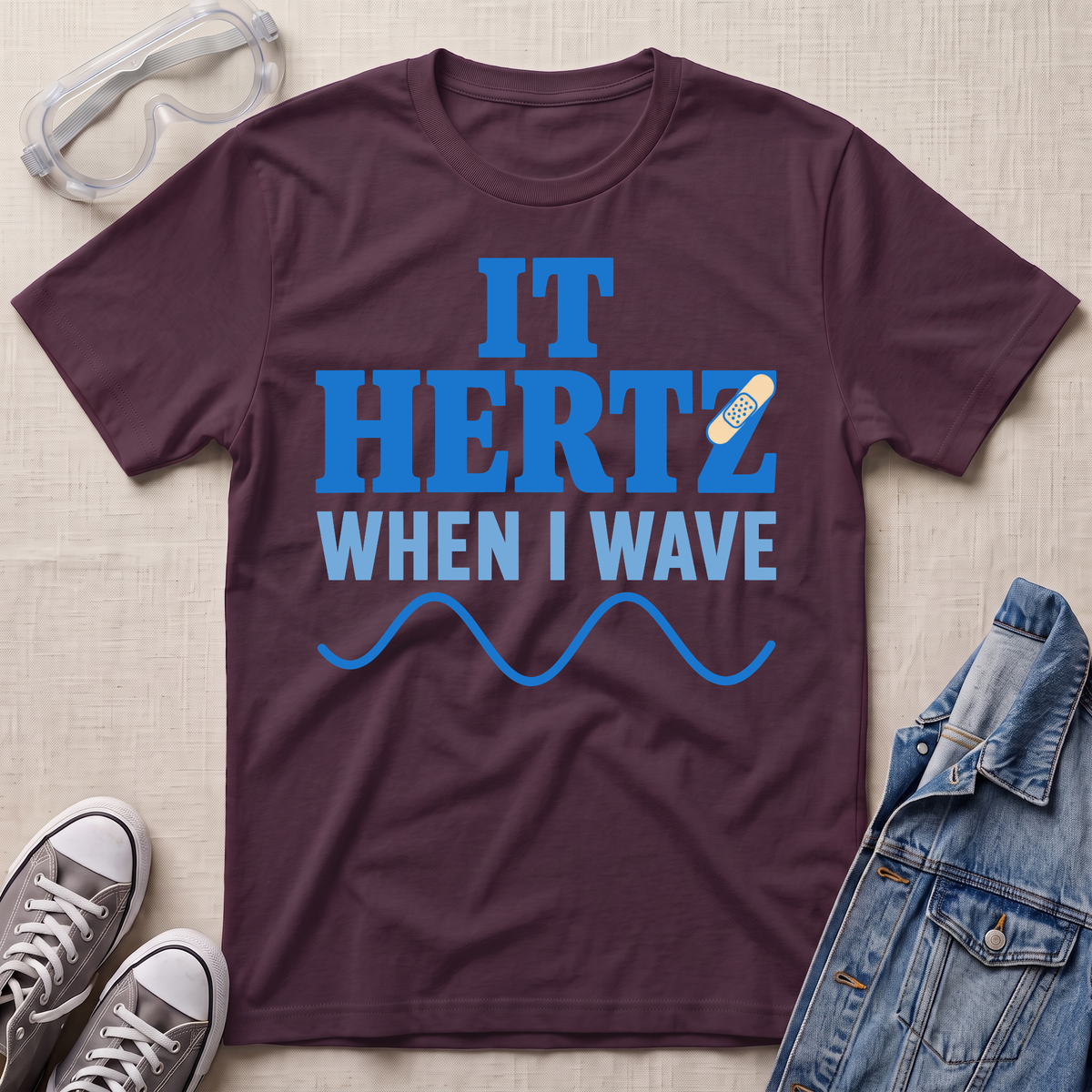 A dark plum purple graphic t-shirt laid flat on a light beige fabric background, surrounded by styling props including clear safety goggles, gray sneakers, and a blue denim jacket. The t-shirt features a blue graphic that reads 'IT HERTZ WHEN I WAVE' with a small band-aid illustration.