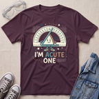 A dark plum purple graphic t-shirt laid flat on a beige fabric background, styled with safety glasses, gray sneakers, and a blue denim jacket. The t-shirt features a cartoon triangle protractor character and the text 'I'M ACUTE ONE'.