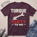 A maroon graphic t-shirt laid flat on a beige fabric background, styled with safety glasses, gray sneakers, and a denim jacket. The t-shirt features a wrench and gear graphic with the text 'TORQUE DIRTY TO ME' in white and red.