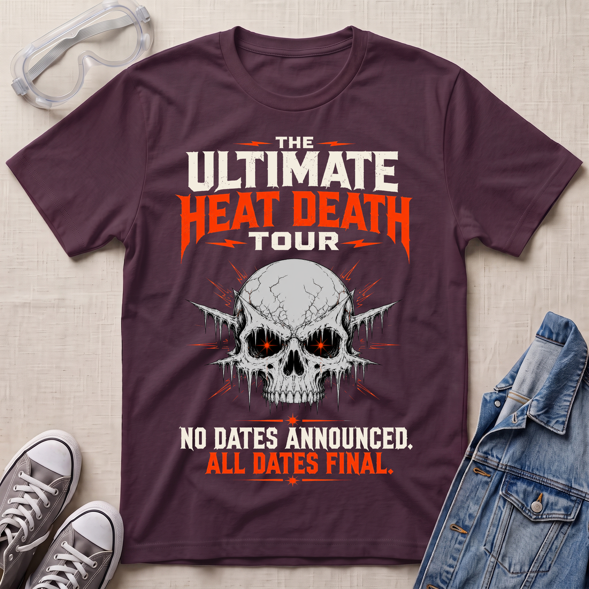 A maroon short-sleeve graphic T-shirt laid flat on a light fabric surface, surrounded by styling props including safety glasses, gray sneakers, and a blue denim jacket. The shirt features a large skull graphic with glowing orange eyes and bold text reading “THE ULTIMATE HEAT DEATH TOUR” and “NO DATES ANNOUNCED.