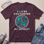 A dark plum purple graphic t-shirt laid flat on a light beige fabric background, styled with safety goggles, gray sneakers, and a denim jacket. The t-shirt features a circular petri-dish style graphic with teal microorganisms and white text that reads 'YOU'RE CULTURING MY INTEREST'.