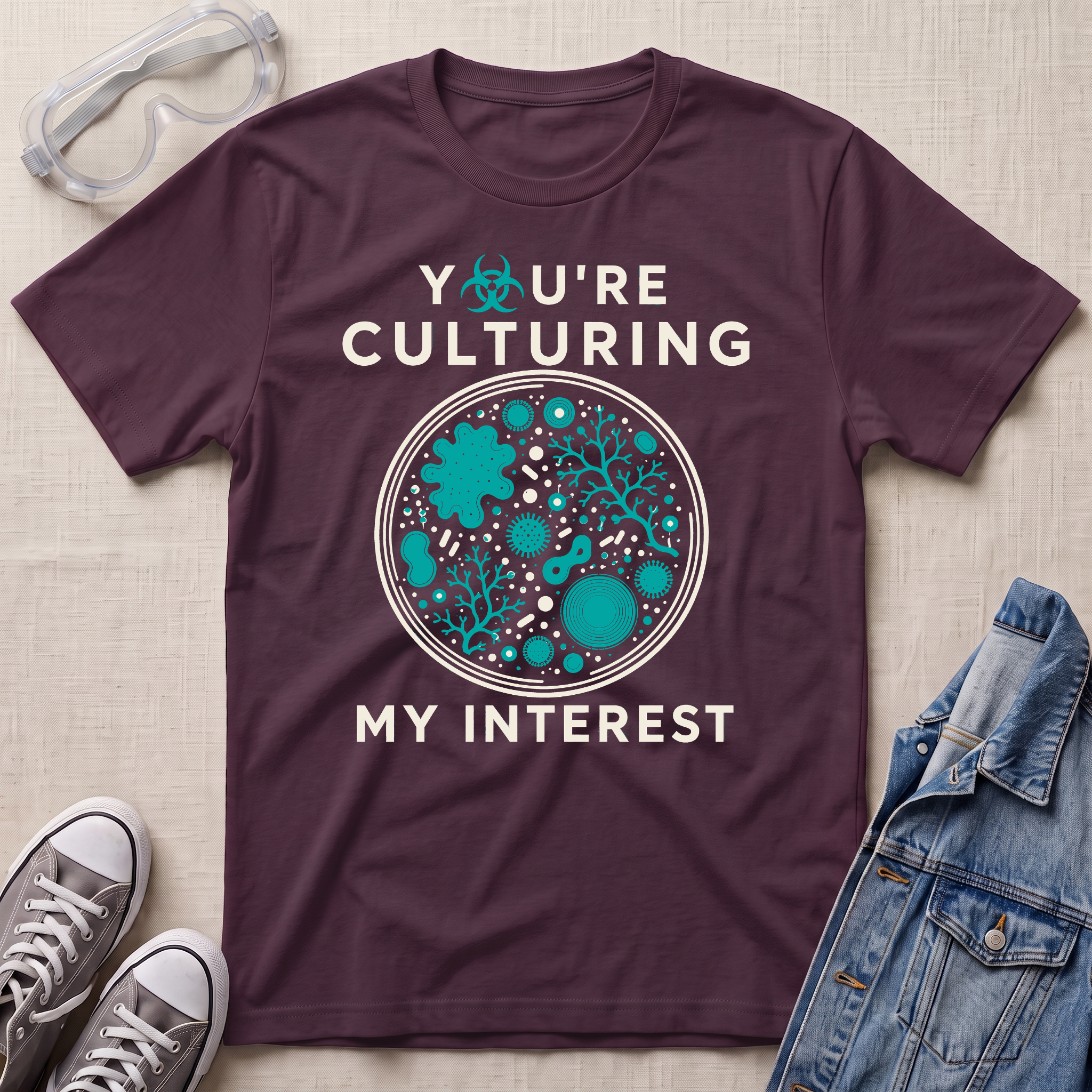 A dark plum purple graphic t-shirt laid flat on a light beige fabric background, styled with safety goggles, gray sneakers, and a denim jacket. The t-shirt features a circular petri-dish style graphic with teal microorganisms and white text that reads 'YOU'RE CULTURING MY INTEREST'.