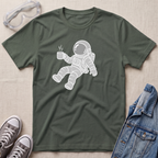 A military green crewneck t-shirt featuring a white line-art graphic of an astronaut floating and holding a steaming mug. The t-shirt is presented in a flat-lay arrangement on a textured beige background, styled with safety goggles, gray sneakers, and a blue denim jacket.
