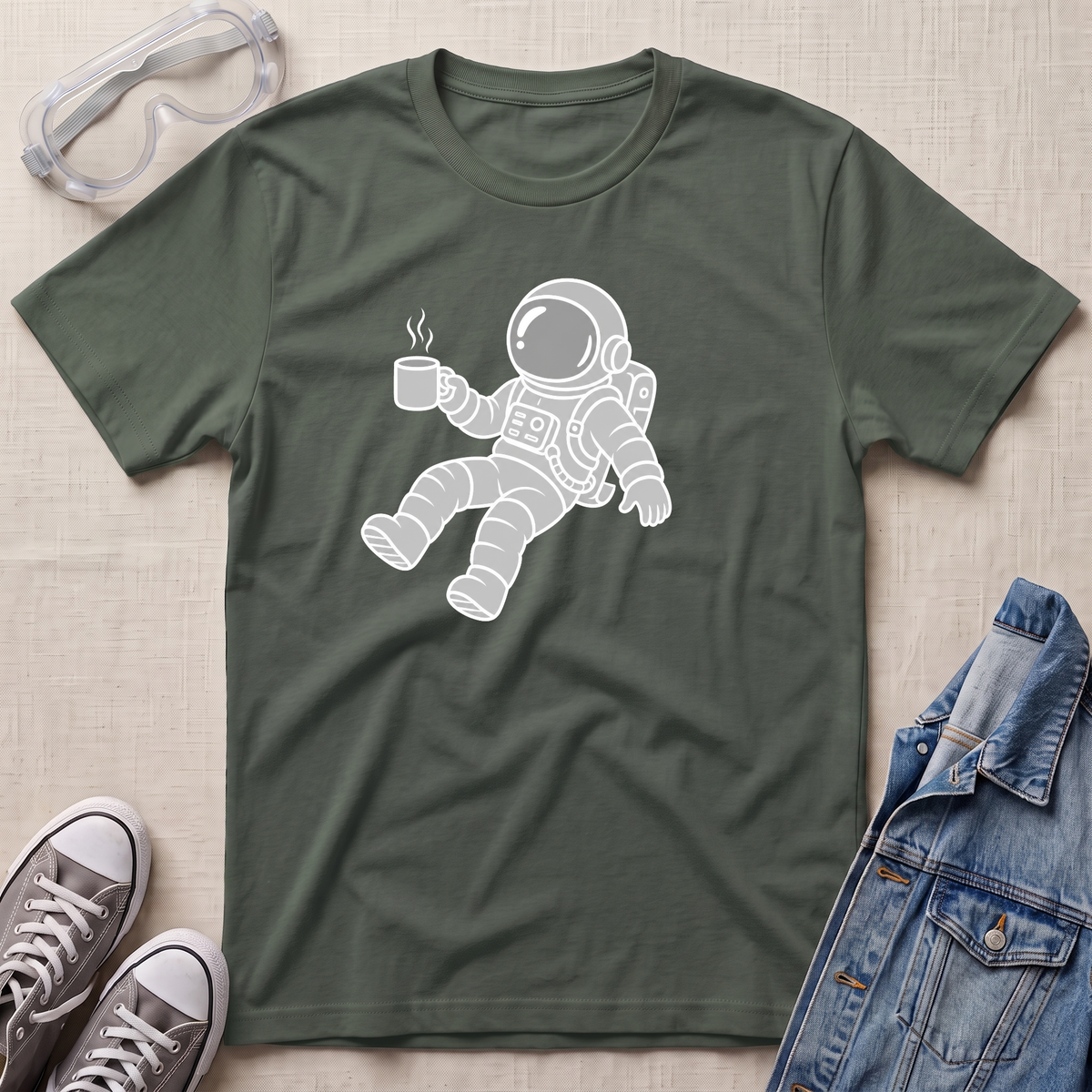 A military green crewneck t-shirt featuring a white line-art graphic of an astronaut floating and holding a steaming mug. The t-shirt is presented in a flat-lay arrangement on a textured beige background, styled with safety goggles, gray sneakers, and a blue denim jacket.