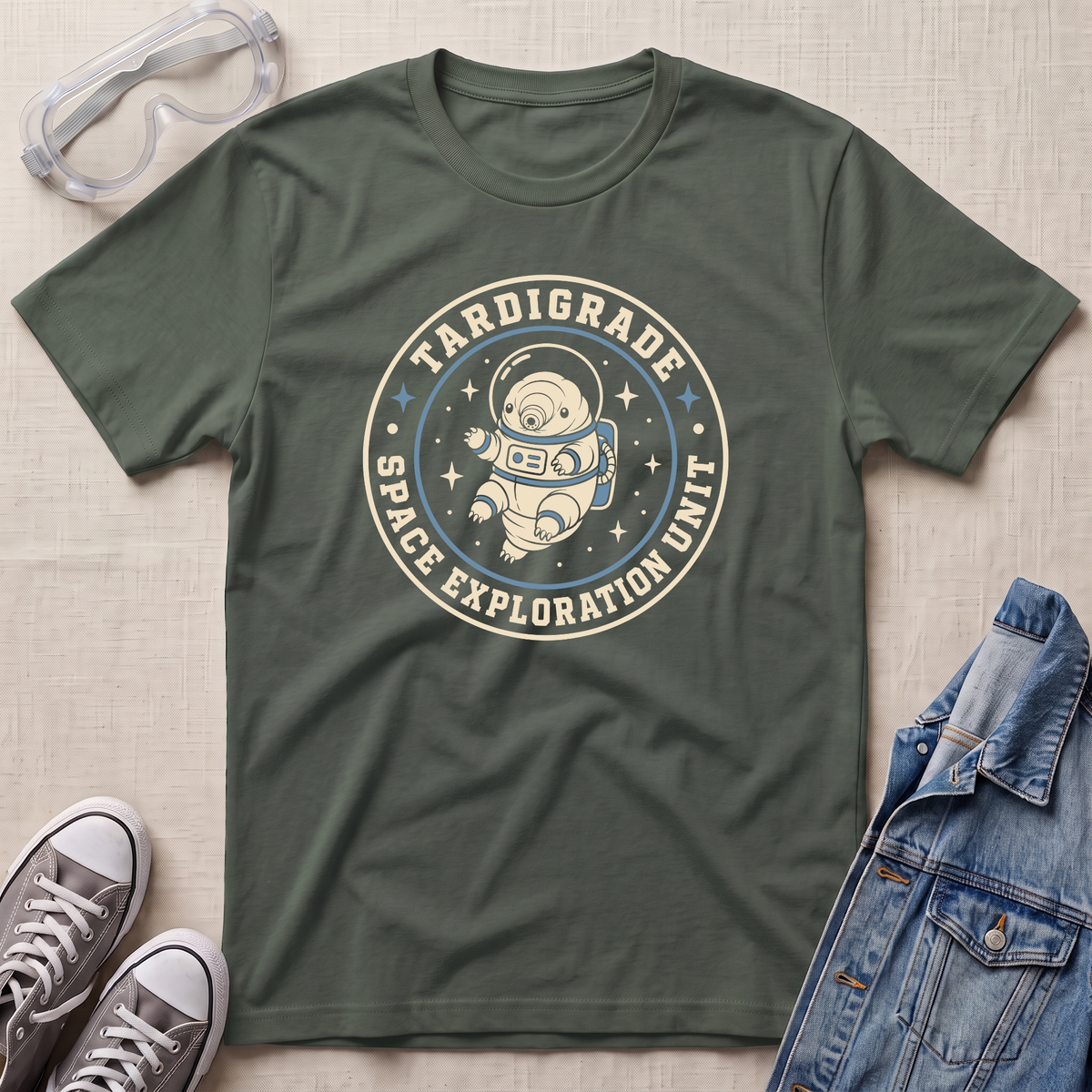 A military green crewneck t-shirt featuring a circular graphic of an astronaut floating in space with the text 'TARDIGRADE SPACE EXPLORATION UNIT'. The shirt is styled in a flat-lay arrangement on a light beige textured background, accompanied by clear safety goggles, gray sneakers, and a blue denim jacket.