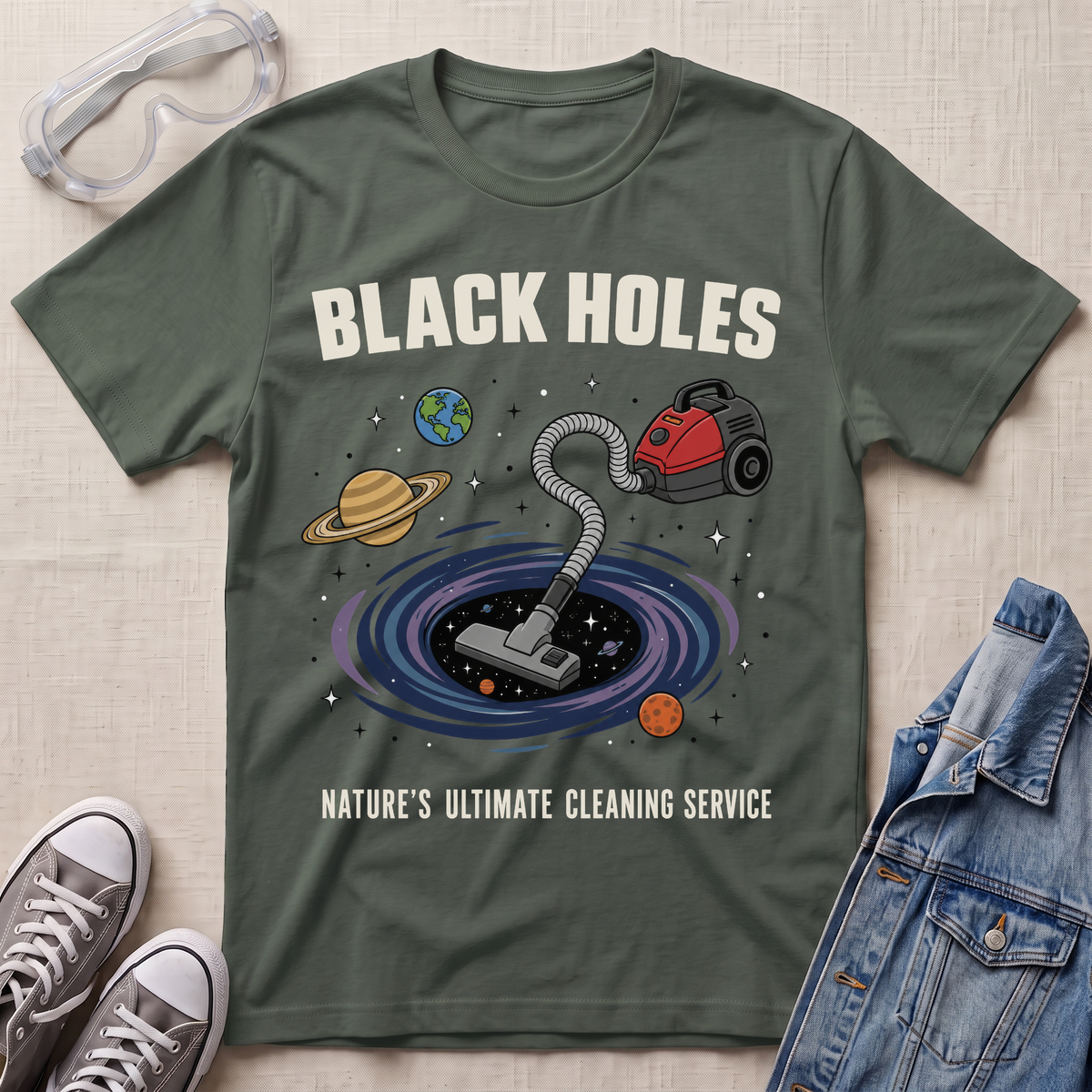 A military green graphic t-shirt laid flat on a textured fabric background, styled with safety goggles, sneakers, and a denim jacket. The t-shirt features a cartoon illustration of a vacuum cleaner falling into a black hole surrounded by planets, with the text 'BLACK HOLES' and 'NATURE'S ULTIMATE CLEANING SERVICE'.
