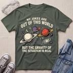 A military green graphic t-shirt laid flat on a light beige textured surface, surrounded by props including clear safety glasses, gray sneakers, and a blue denim jacket. The t-shirt features a space-themed graphic with planets, stars, and the text 'MY JOKES ARE OUT OF THIS WORLD BUT THE GRAVITY OF THE SITUATION IS REAL'.