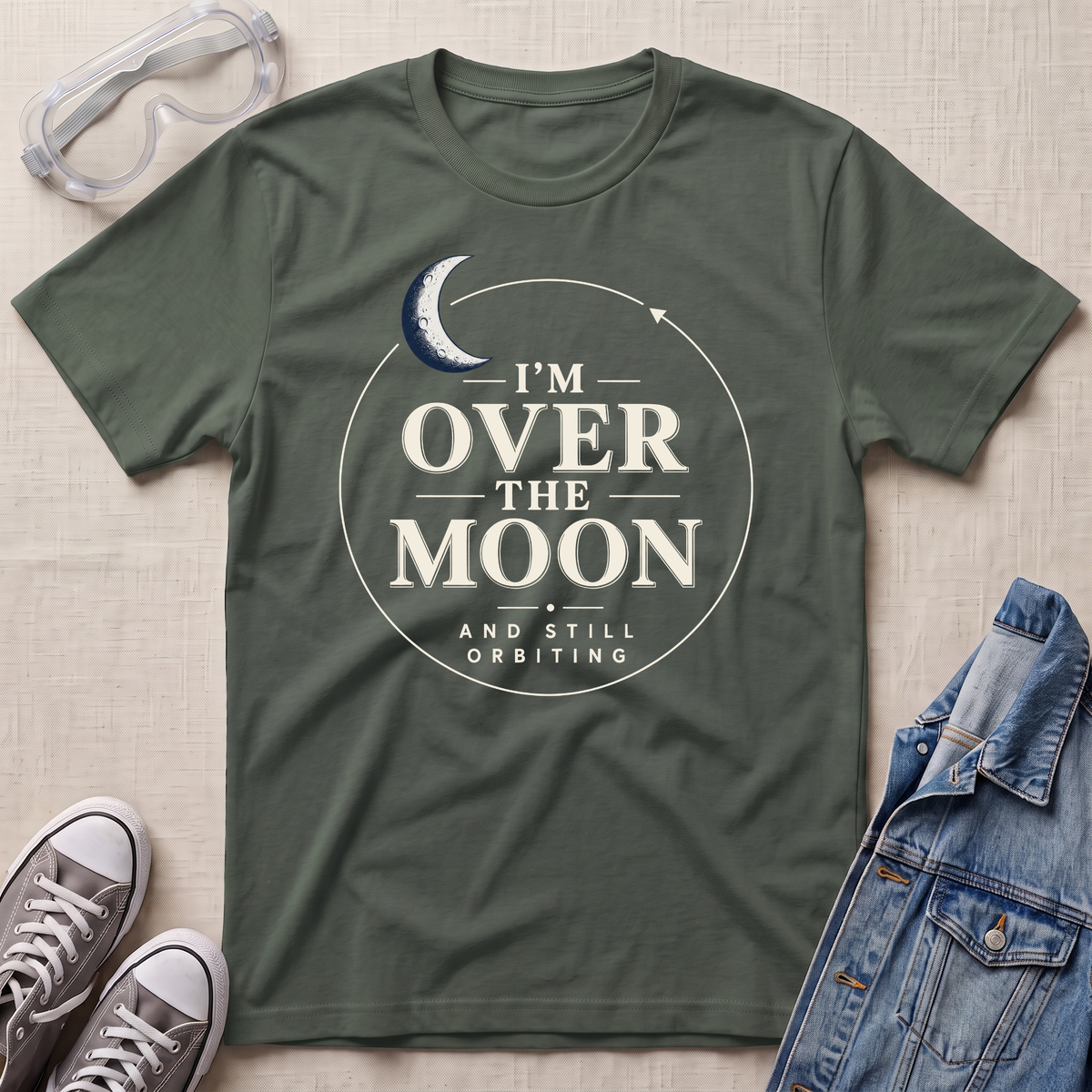 A military green graphic t-shirt laid flat on a textured beige fabric background, styled with clear safety goggles, gray sneakers, and a blue denim jacket. The t-shirt features a white circular graphic with a crescent moon and the text 'I'M OVER THE MOON AND STILL ORBITING'.