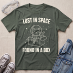 An military green graphic t-shirt laid flat on a light beige textured background, surrounded by styling props including safety goggles, gray sneakers, and a denim jacket. The t-shirt features a white graphic of a cat in a space helmet sitting in a box with the text 'LOST IN SPACE' and 'FOUND IN A BOX'.