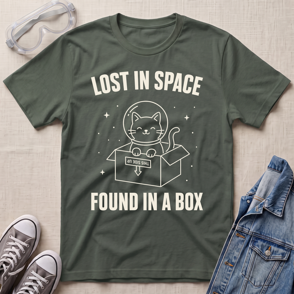 An military green graphic t-shirt laid flat on a light beige textured background, surrounded by styling props including safety goggles, gray sneakers, and a denim jacket. The t-shirt features a white graphic of a cat in a space helmet sitting in a box with the text 'LOST IN SPACE' and 'FOUND IN A BOX'.