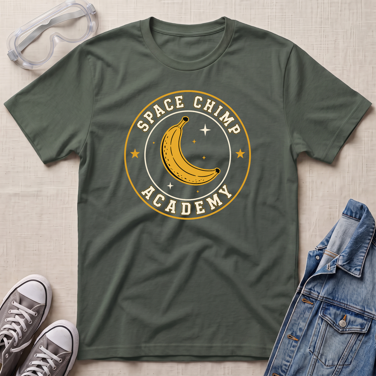 A military green t-shirt laid flat on a textured beige fabric background, styled with a 'Space Chimp Academy' banana graphic. Surrounding props include clear safety goggles, gray sneakers, and a blue denim jacket.