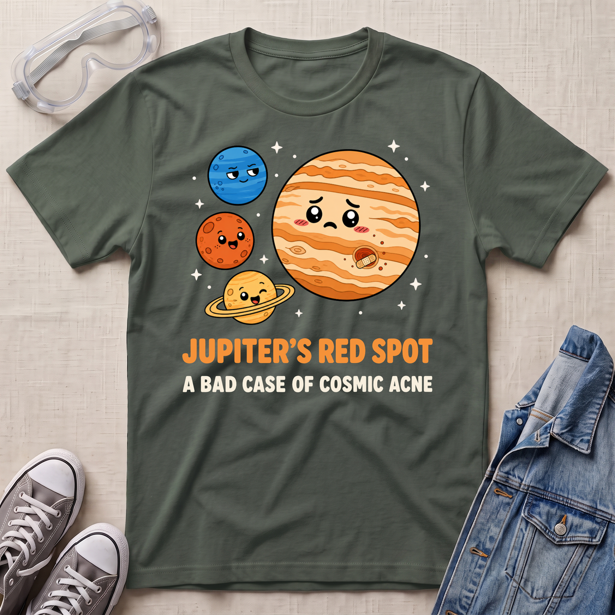An military green graphic t-shirt laid flat on a light beige textured background, styled with safety glasses, gray sneakers, and a denim jacket. The t-shirt features a cartoon graphic of Jupiter, Mars, and Saturn with the text 'JUPITER'S RED_spot A BAD CASE OF COSMIC ACNE'.
