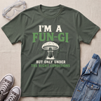 A military green graphic t-shirt laid flat on a light beige textured background, styled with safety goggles, gray sneakers, and a denim jacket. The t-shirt features a mushroom graphic and the text 'I'M A FUN-GI BUT ONLY UNDER THE RIGHT CONDITIONS'.