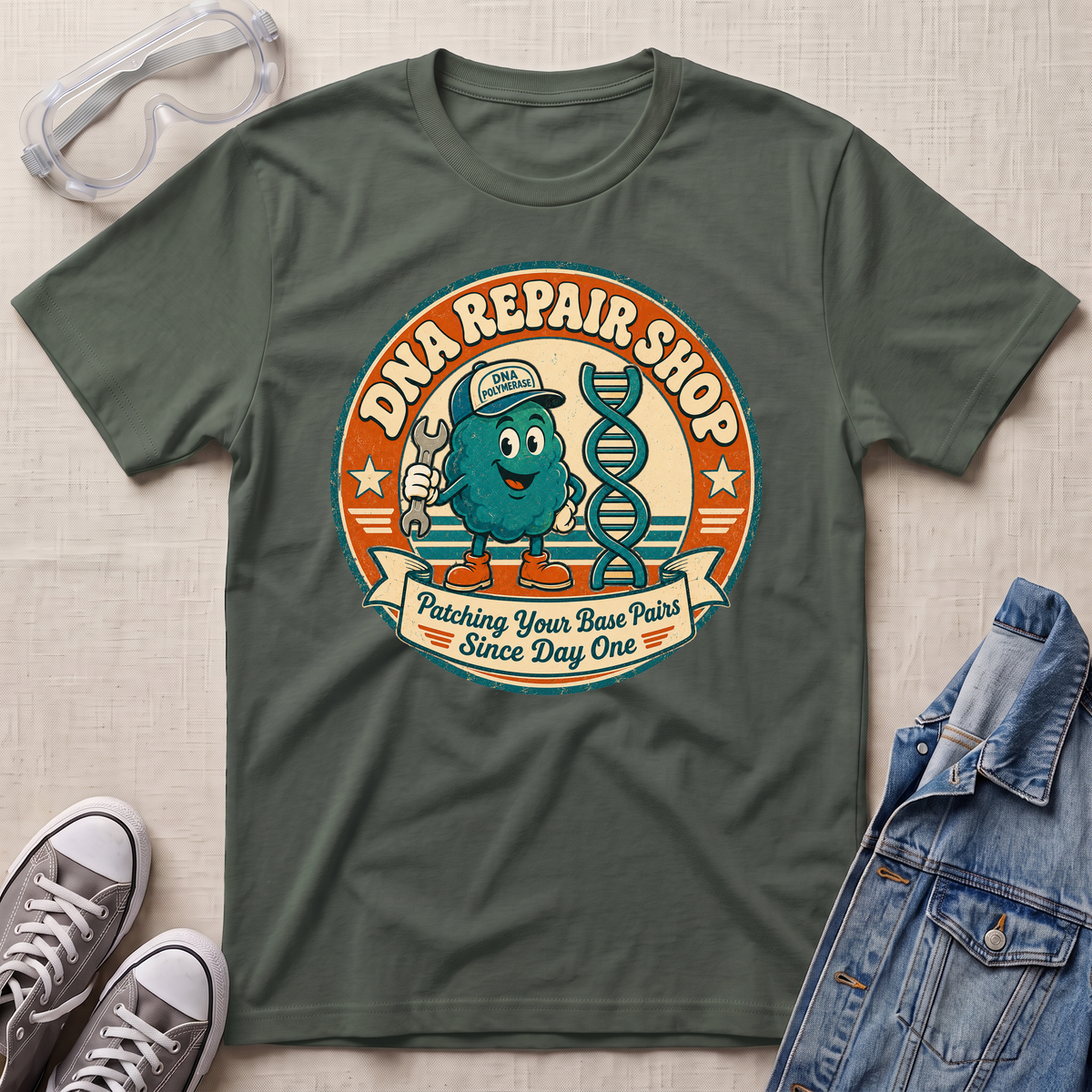 An olive green graphic t-shirt laid flat on a textured beige surface, surrounded by styling props including safety goggles, gray sneakers, and a denim jacket. The t-shirt features a retro circular graphic for 'DNA REPAIR SHOP' with a cartoon teal character and a DNA double helix.