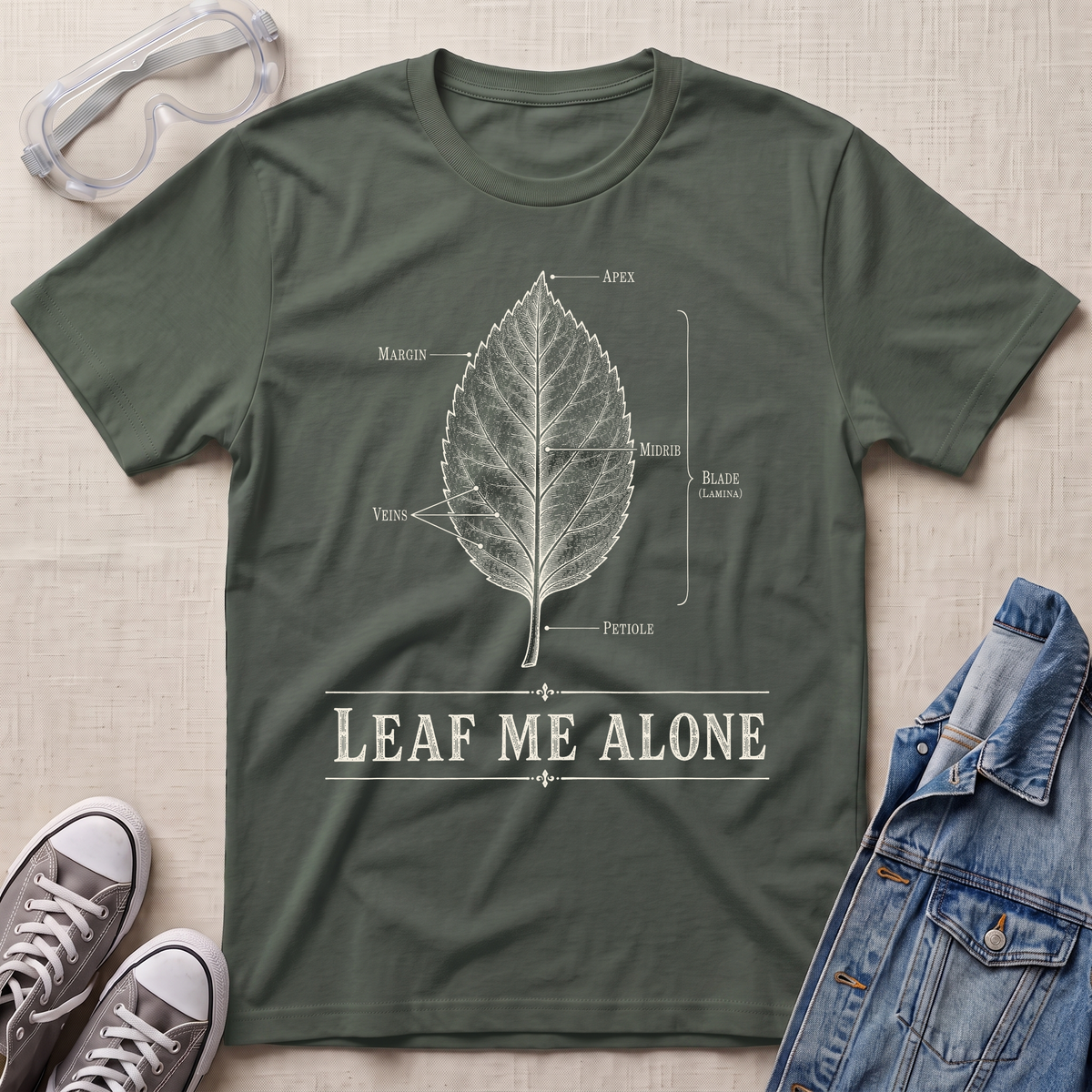 A military green botanical t-shirt featuring a vintage labeled leaf anatomy illustration with the pun “Leaf Me Alone” in bold retro typography.