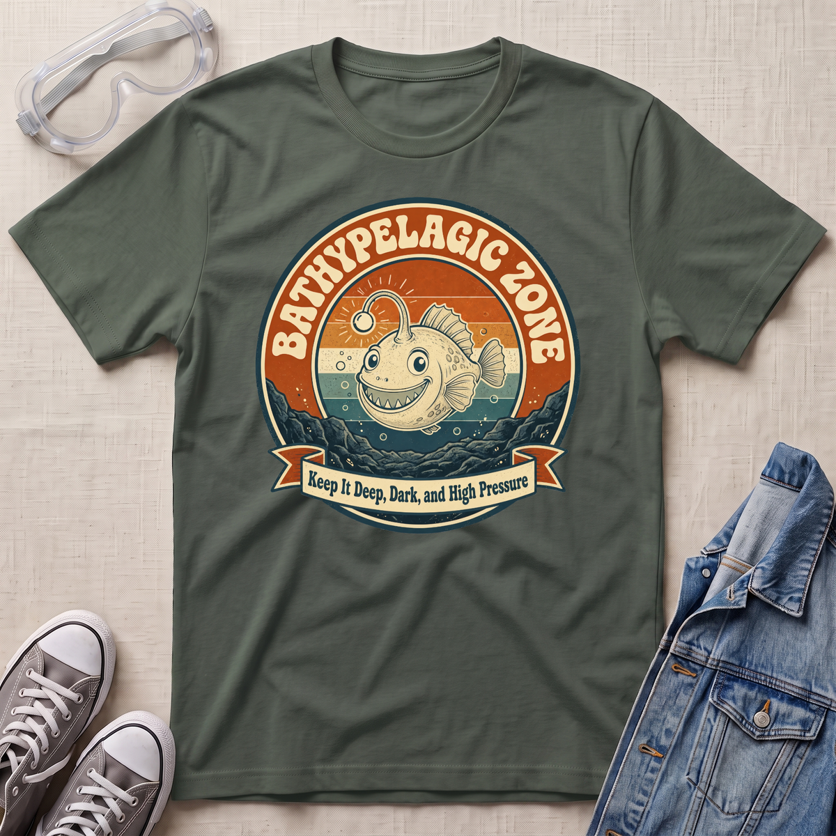 An olive green graphic t-shirt laid flat on a textured beige surface, styled with safety goggles, gray sneakers, and a denim jacket. The t-shirt features a retro circular graphic of a smiling fish in deep water with the text 'BATHYPELAGIC ZONE' and 'Keep it Deep, Dark, and High Pressure'.