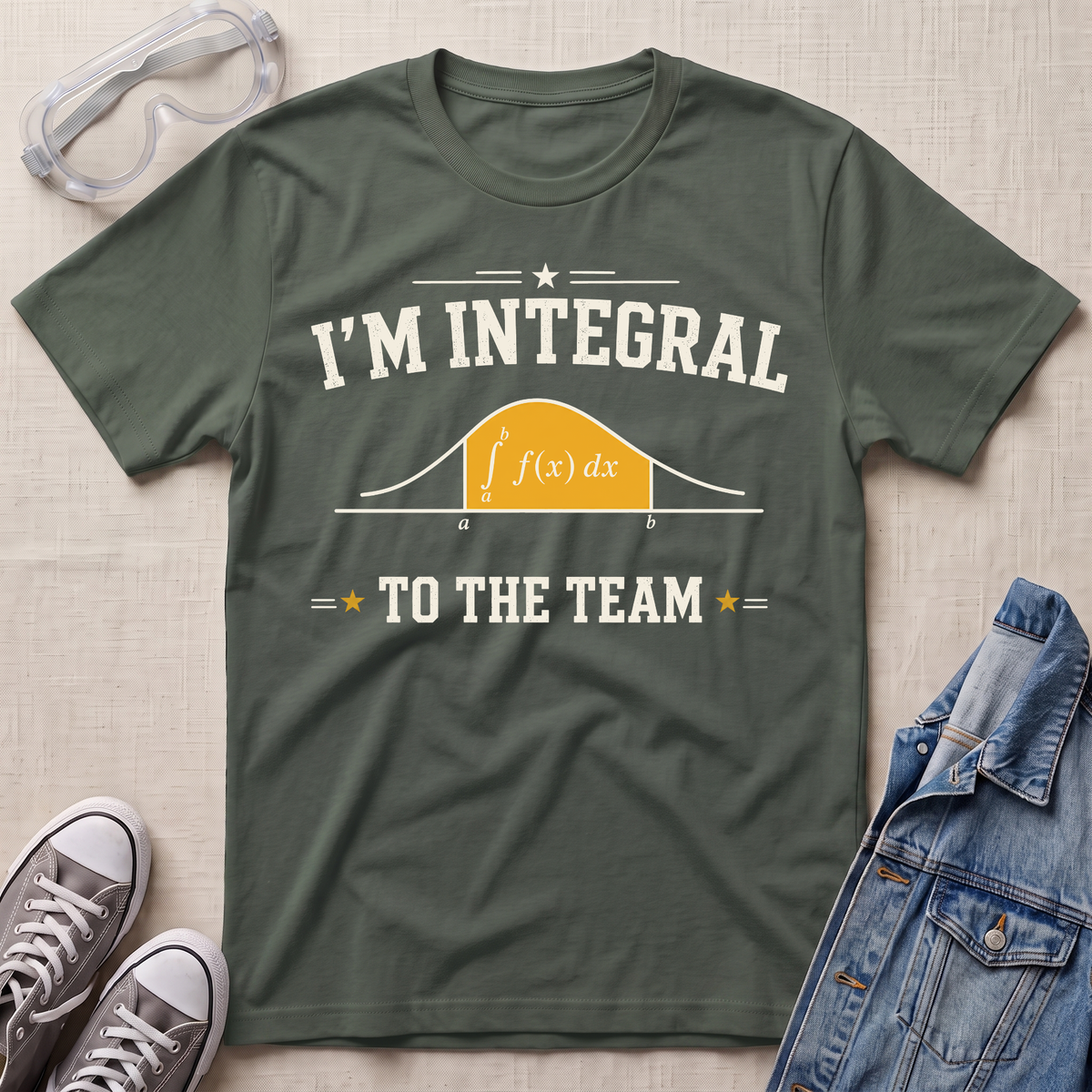 An olive green graphic t-shirt laid flat on a textured beige background, surrounded by styling props including safety glasses, gray sneakers, and a blue denim jacket. The t-shirt features a white and orange graphic that reads 'I'M INTEGRAL TO THE TEAM' with a simple integral curve.