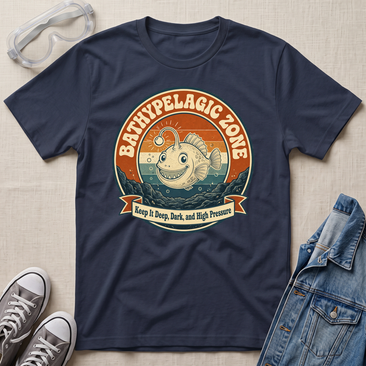A navy blue graphic t-shirt laid flat on a beige fabric background, styled with safety goggles, gray sneakers, and a denim jacket. The t-shirt features a circular retro-style graphic of a smiling fish in the bathypelagic zone with the text 'BATHYPELAGIC ZONE' and 'Keep it Deep, Dark, and High Pressure'.