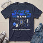 A navy blue graphic t-shirt featuring a 'Quantum Mechanics is Easy' design in blue and white, styled in a flat-lay arrangement with clear safety goggles, gray sneakers, and a denim jacket on a beige fabric background.