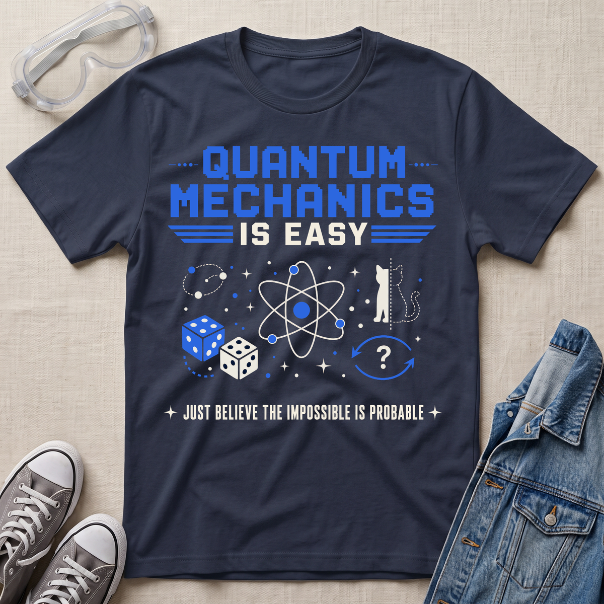 A navy blue graphic t-shirt featuring a 'Quantum Mechanics is Easy' design in blue and white, styled in a flat-lay arrangement with clear safety goggles, gray sneakers, and a denim jacket on a beige fabric background.