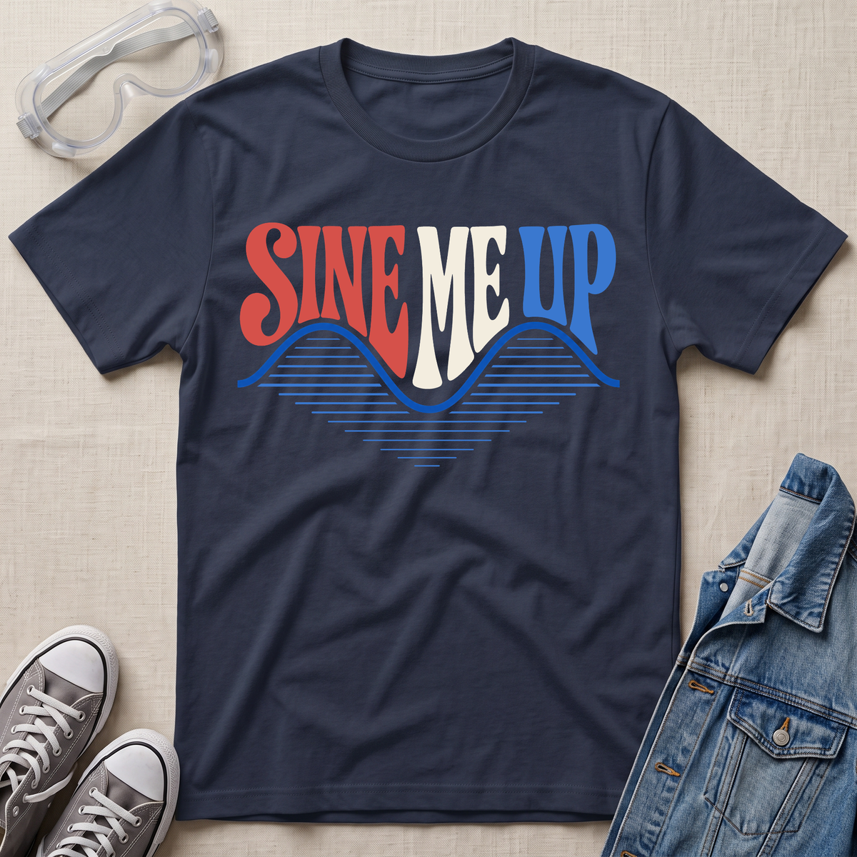 A navy blue graphic t-shirt laid flat on a textured beige fabric background, styled with safety goggles, gray sneakers, and a denim jacket. The t-shirt features a retro-style graphic reading 'SINE ME UP' in red, white, and blue, with a blue soundwave design underneath.
