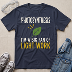 A navy blue graphic t-shirt laid flat on a beige fabric background, surrounded by styled props including safety goggles, gray sneakers, and a denim jacket. The t-shirt features a green leaf graphic and the text 'PHOTOSYNTHESIS I'M A BIG FAN OF LIGHT WORK' in white and yellow.
