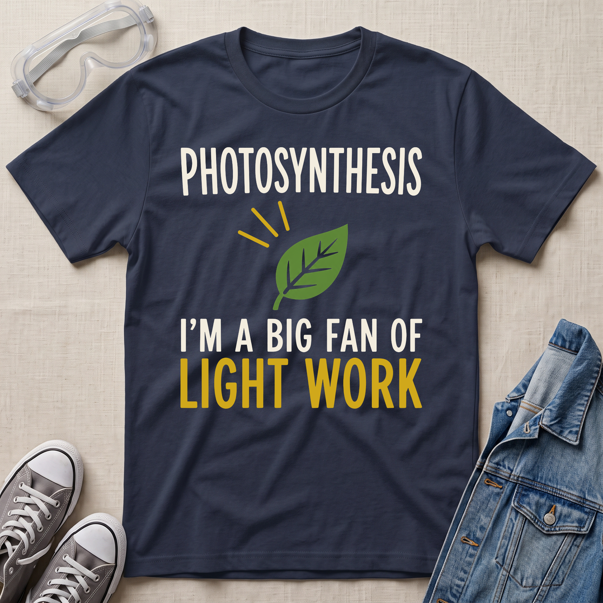 A navy blue graphic t-shirt laid flat on a beige fabric background, surrounded by styled props including safety goggles, gray sneakers, and a denim jacket. The t-shirt features a green leaf graphic and the text 'PHOTOSYNTHESIS I'M A BIG FAN OF LIGHT WORK' in white and yellow.