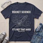 A navy blue graphic t-shirt featuring a white rocket science blueprint design with the text 'ROCKET SCIENCE IT'S NOT THAT HARD (Wait, yes it is.)' laid flat on a beige fabric background, styled with safety goggles, sneakers, and a denim jacket.