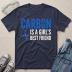 A navy blue graphic t-shirt laid flat on a beige fabric background, surrounded by styling props including safety goggles, gray sneakers, and a denim jacket. The t-shirt features the text 'CARBON IS A GIRL'S BEST FRIEND' in blue and white, with a small blue molecule graphic.