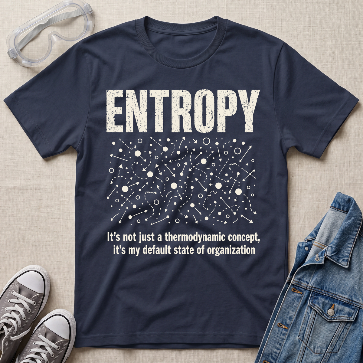 A navy blue graphic t-shirt laid flat on a beige fabric background, styled with safety goggles, gray sneakers, and a denim jacket. The t-shirt features a white distressed graphic of the word 'ENTROPY' above a entropy pattern, with the text 'It's not just a thermodynamic concept, it's my default state of organization' below.
